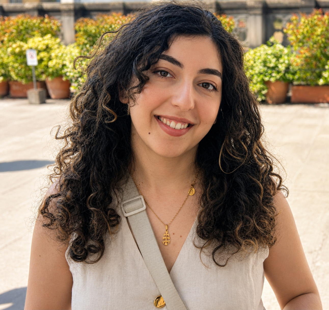 Smiling person with curly hair outdoors, wearing a light sleeveless top and necklace.