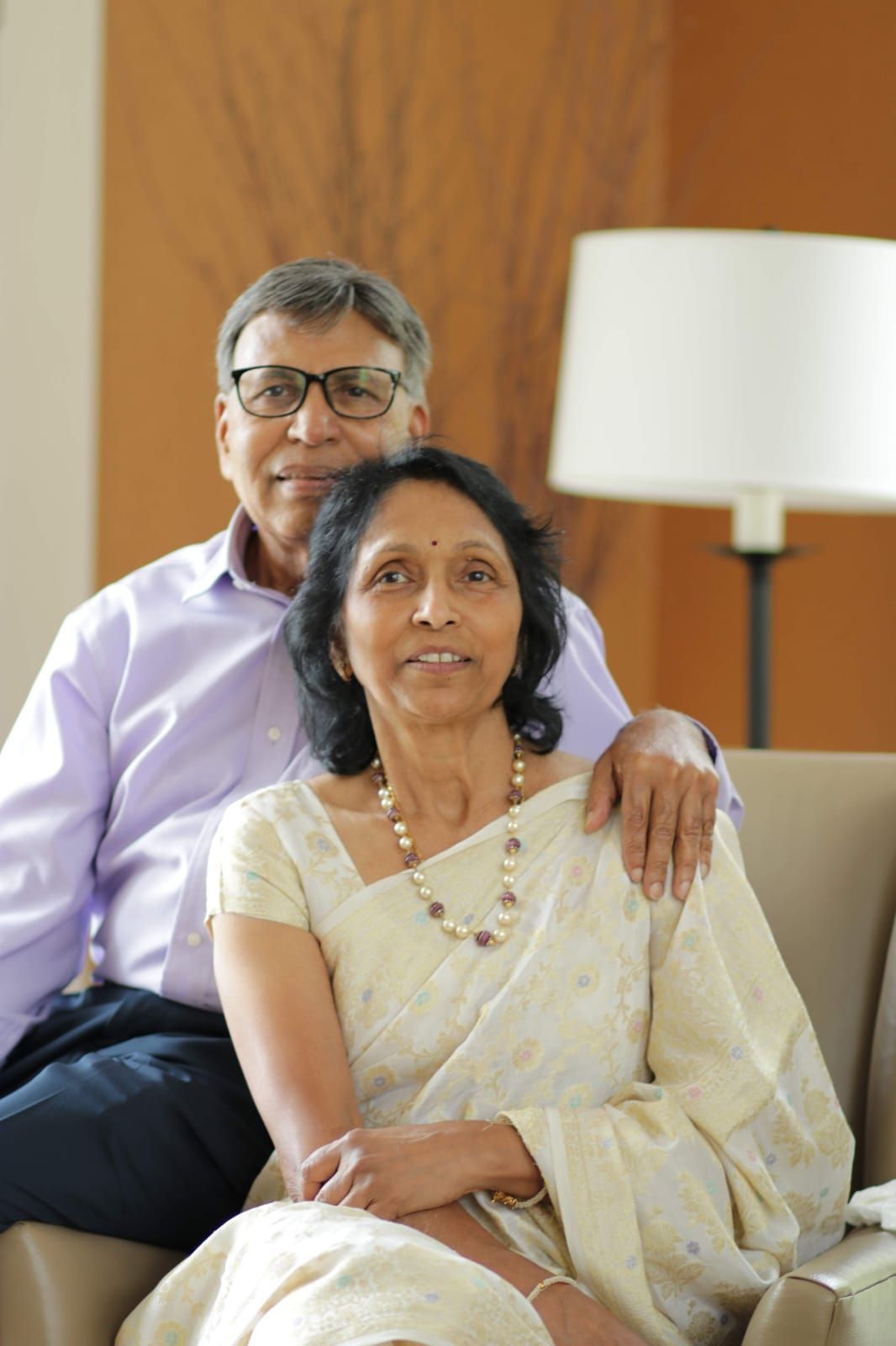 Couple seated on a sofa, with the woman in a cream saree in front and a lamp in the background.