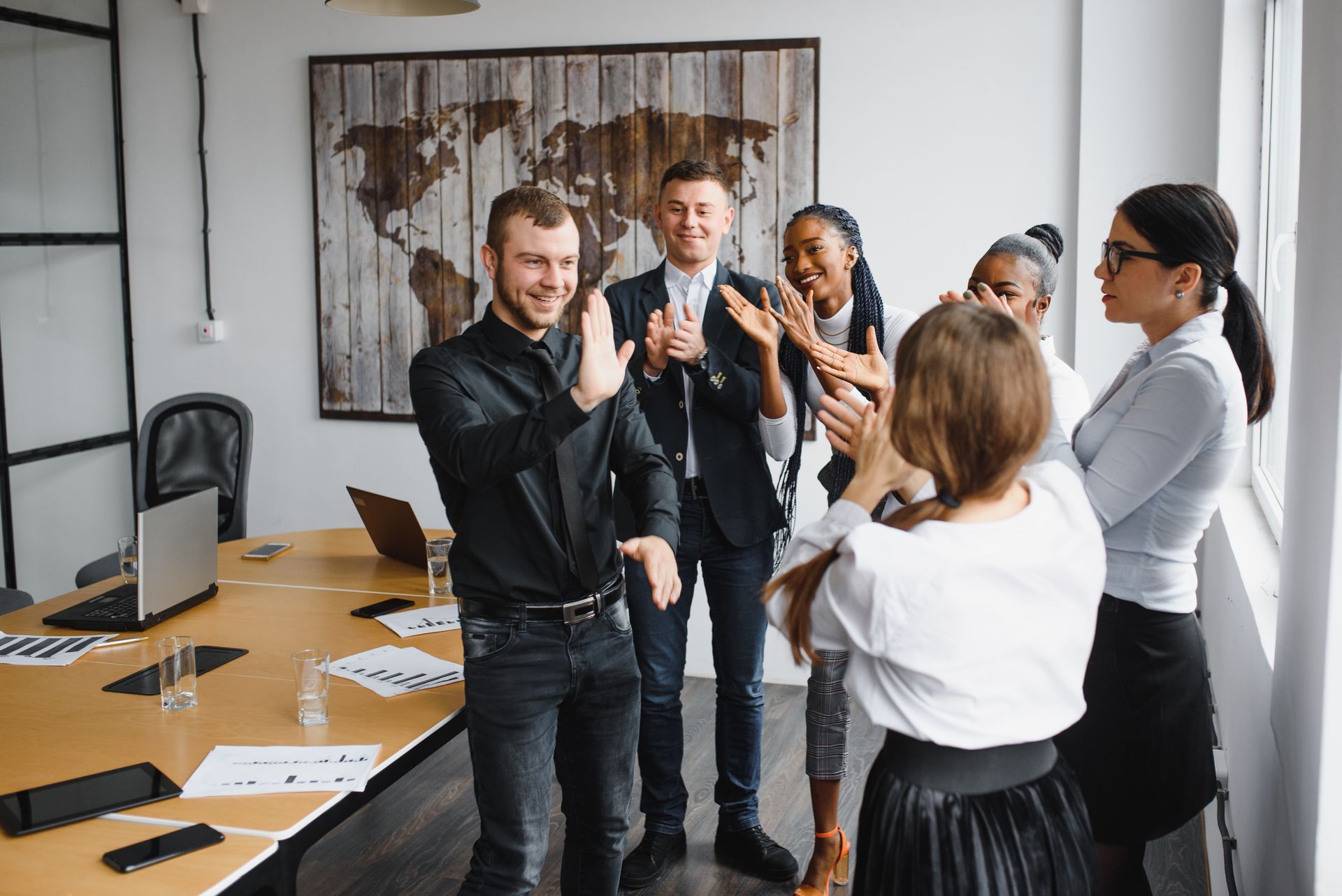 Colleagues in business attire clapping in an office meeting room by a desk and wall art