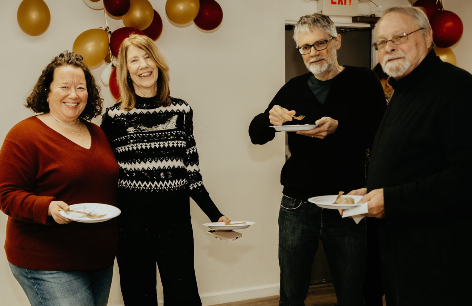 Four people holding plates pose at a party with balloons on the wall.