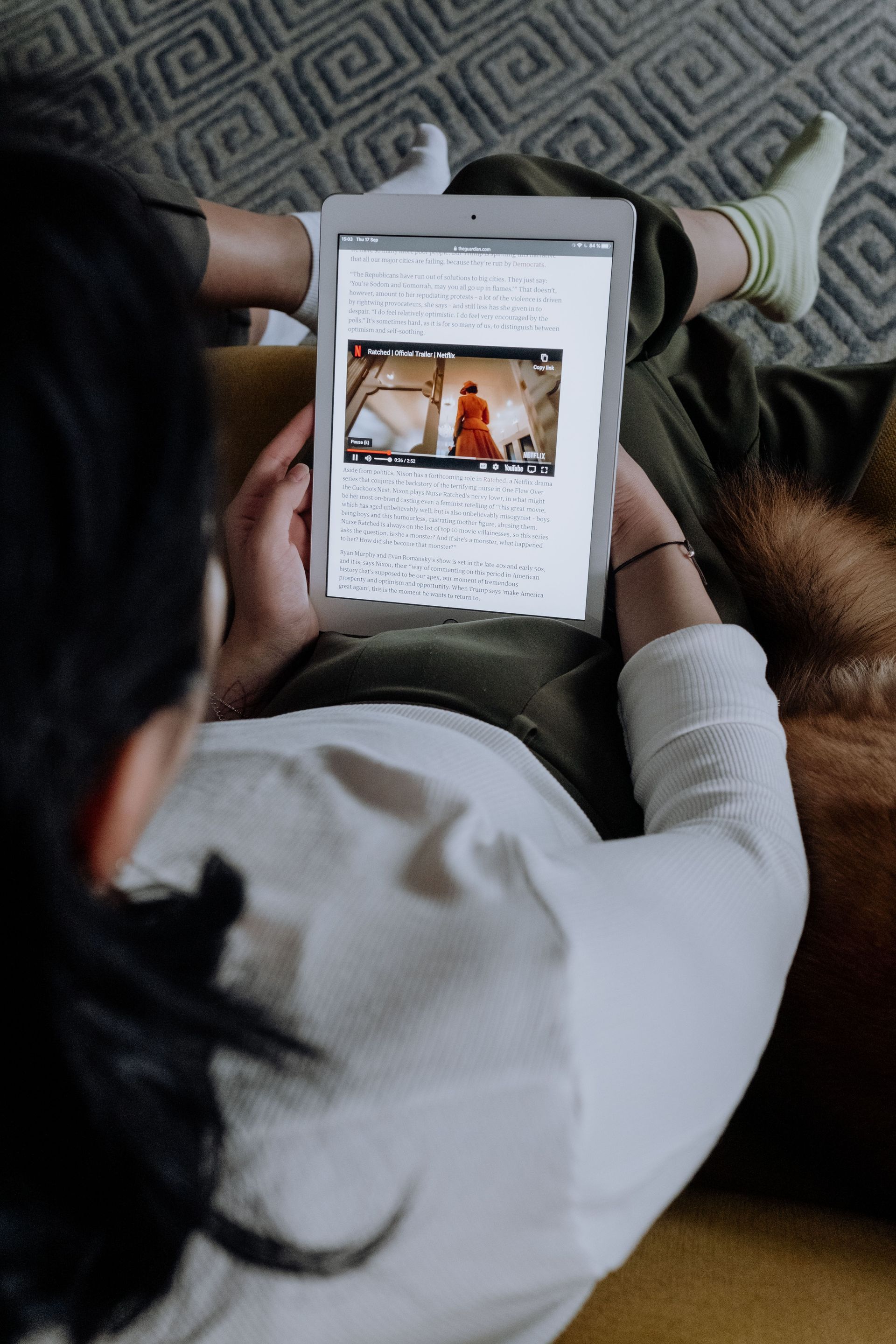Person holding a tablet while seated on a couch, viewing a video with a small child onscreen.