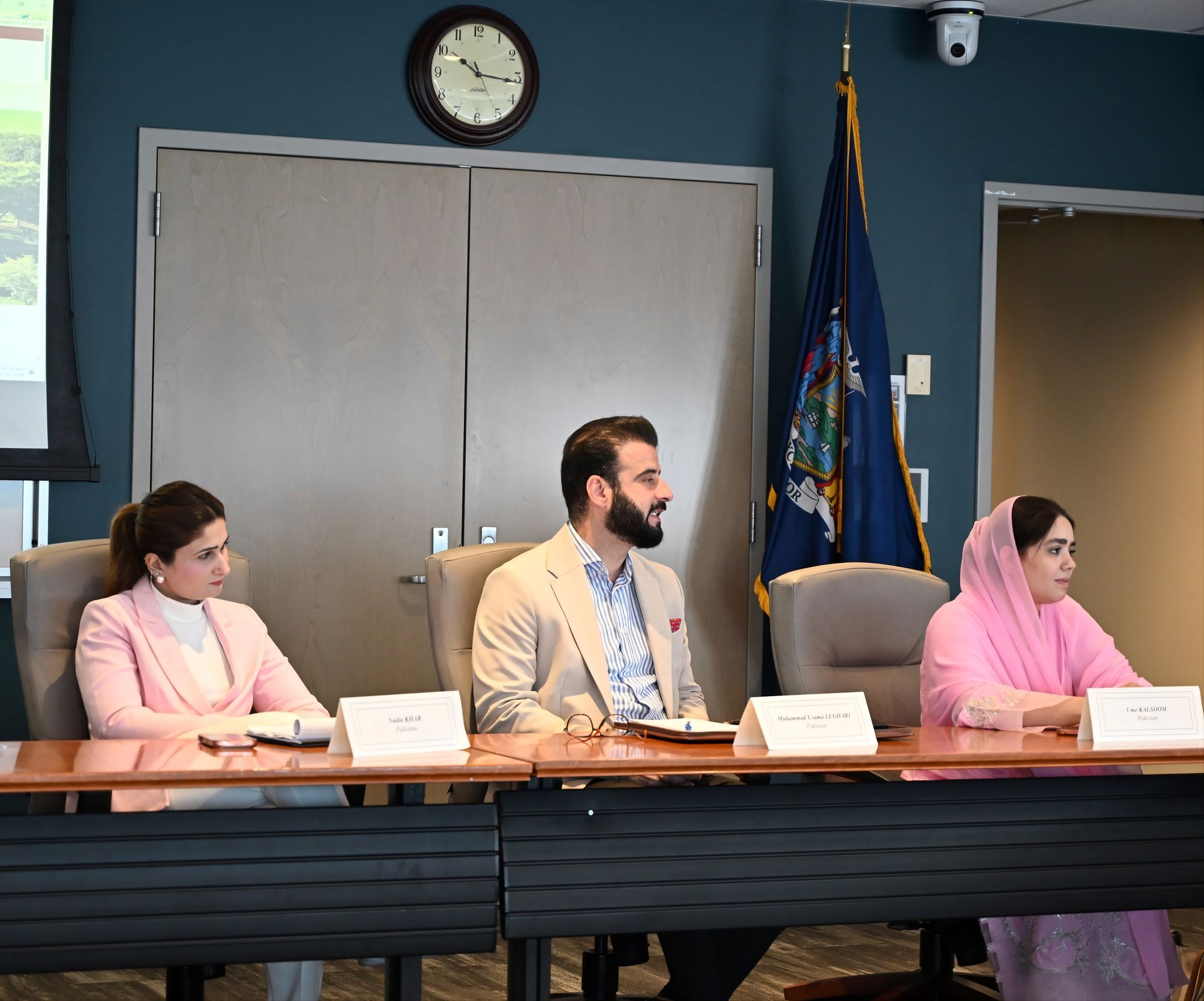 Three people seated at a conference table in a meeting room, with name placards and a clock on the wall.
