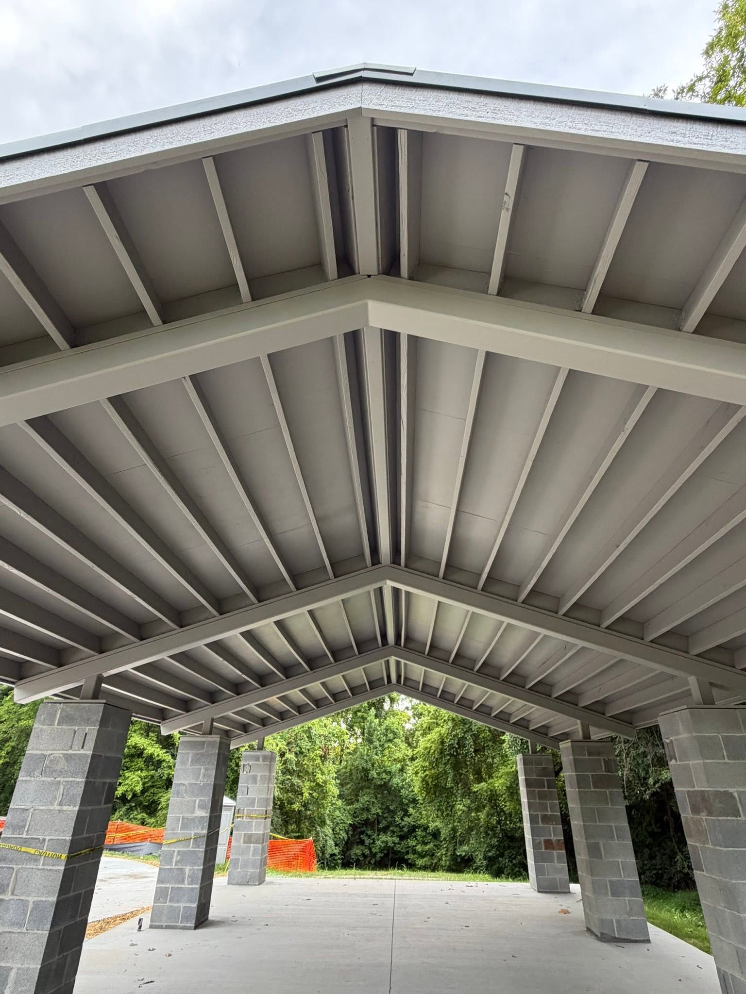 Covered pavilion with stone columns and ribbed metal roof, opening onto a tree-lined outdoor area.