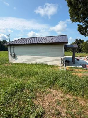 White lakeside shed with dark roof beside a dock, under a blue sky and grassy shoreline.