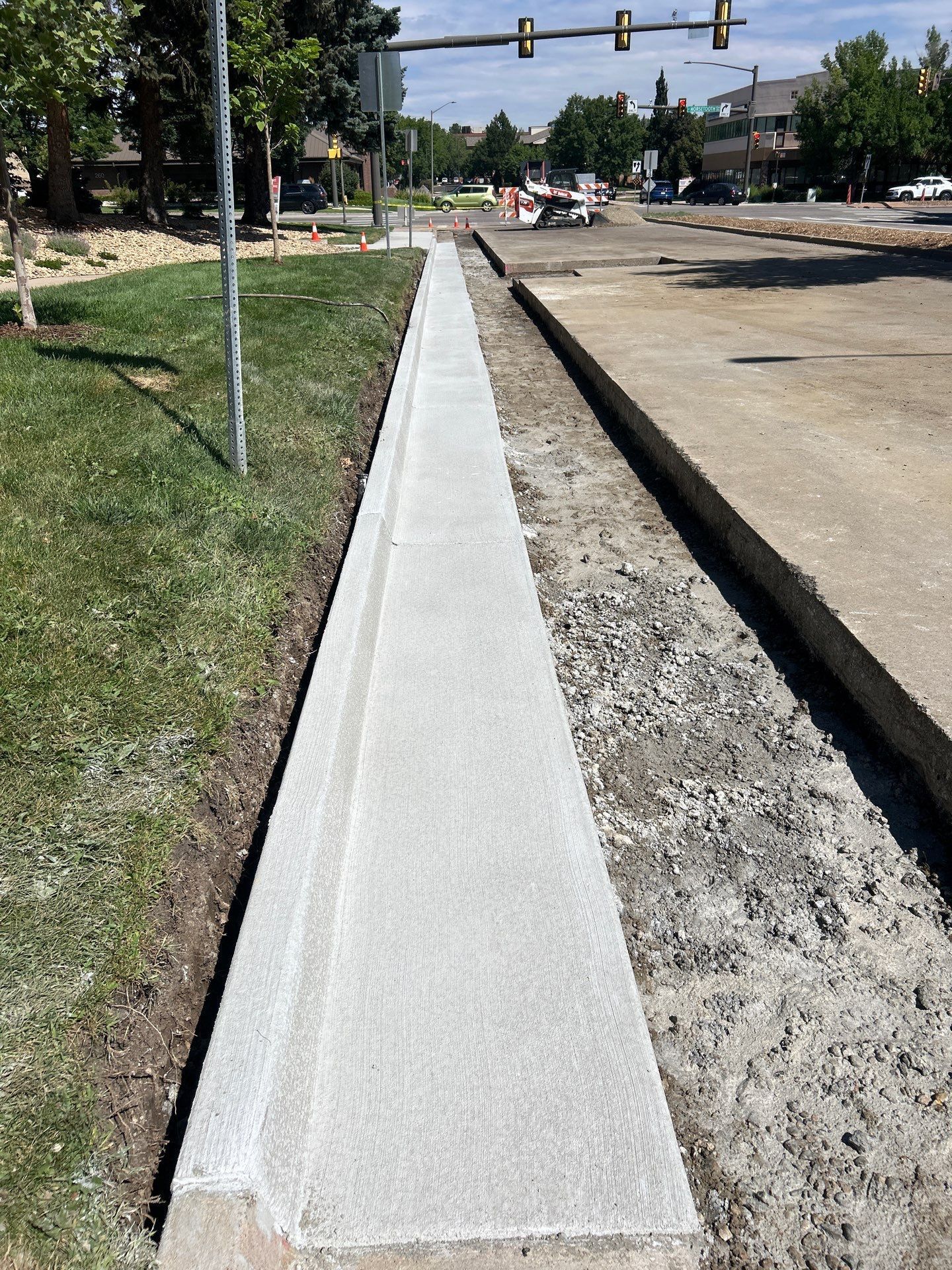 New concrete sidewalk next to grass and road under construction; traffic lights in the distance.