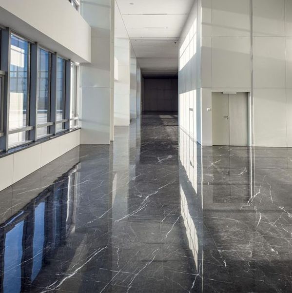Long, bright hallway with reflective, dark stone floor, tall windows, and white walls.