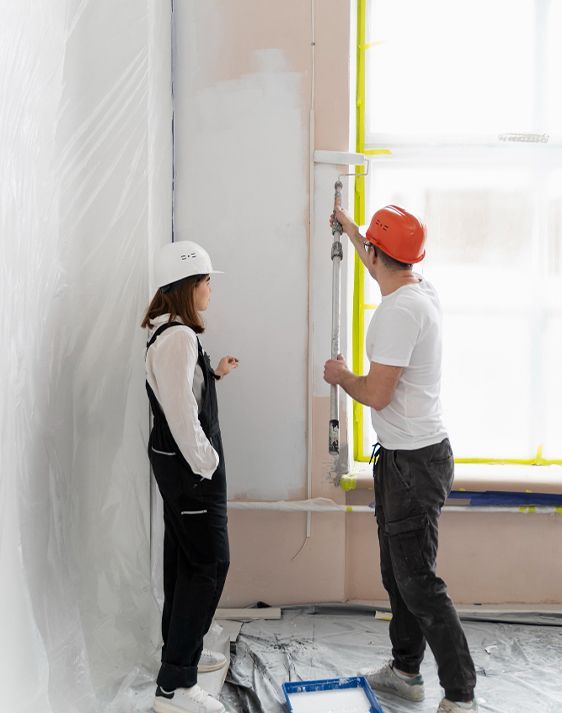 Two people, wearing hard hats, painting a wall. One is using a roller, the other is observing.