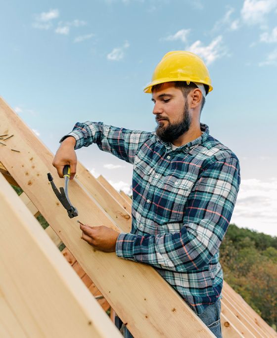 Construction worker hammering a nail onto a wooden roof frame, wearing a yellow hard hat and a plaid shirt.
