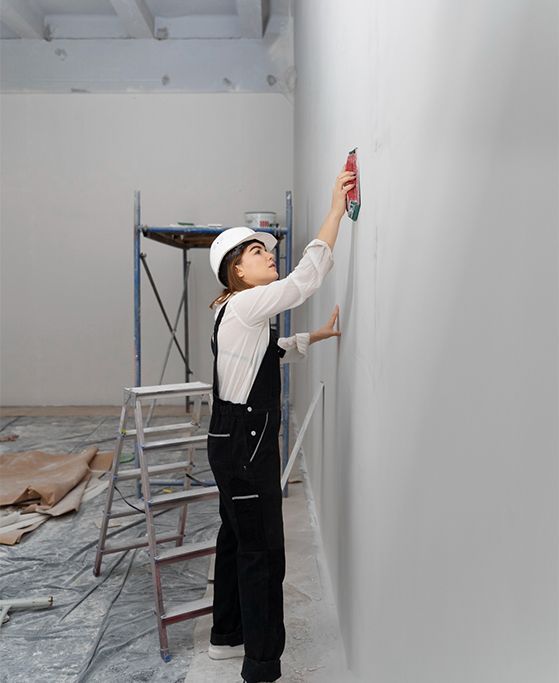 Woman in a hard hat and overalls measuring a wall in a room under construction.