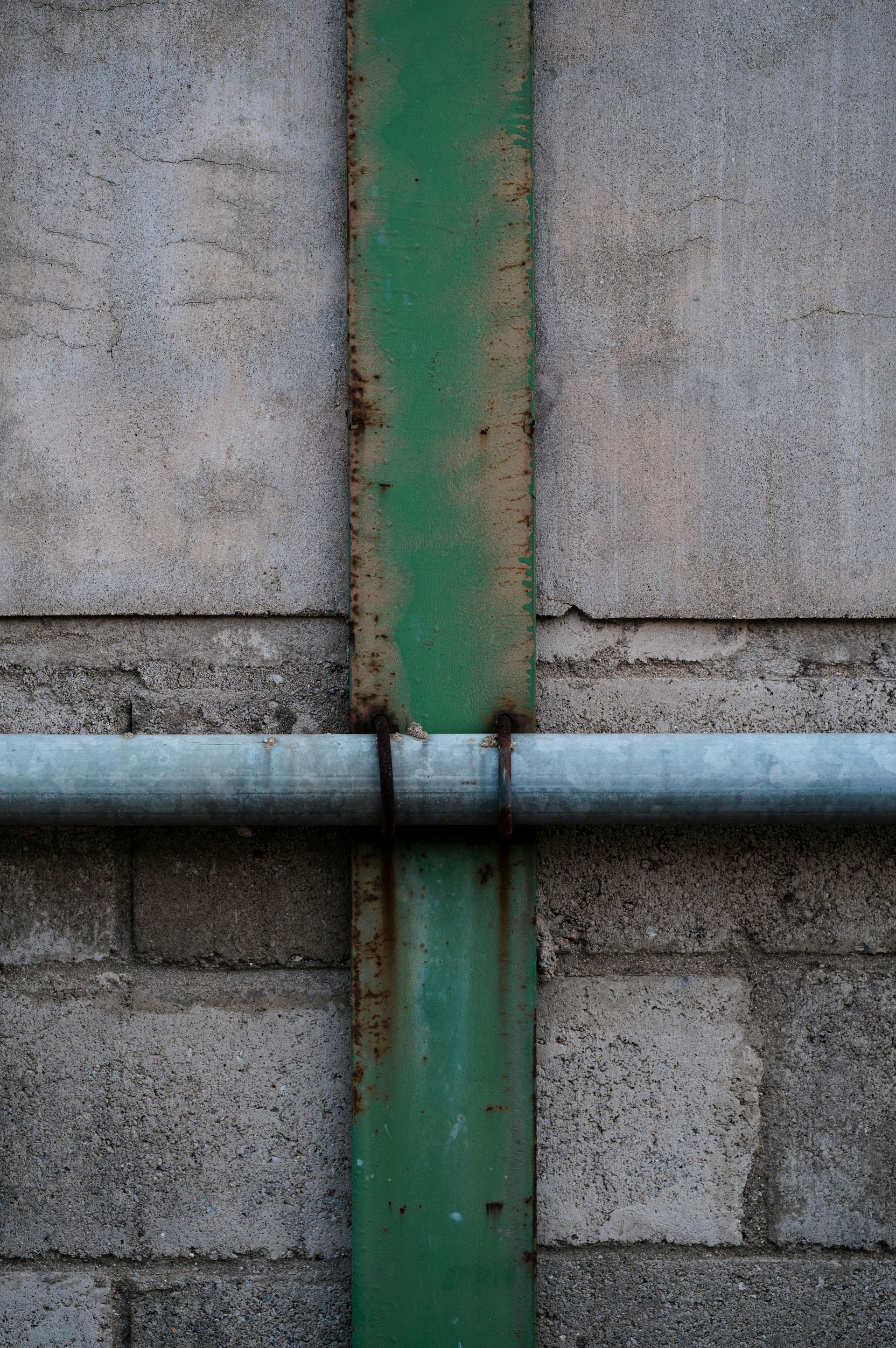 Green metal beam crosses a gray pipe on a concrete block wall.