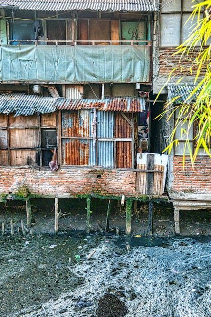 Dilapidated buildings on stilts over murky water. Rusting metal, weathered wood, and a pale blue tarp.