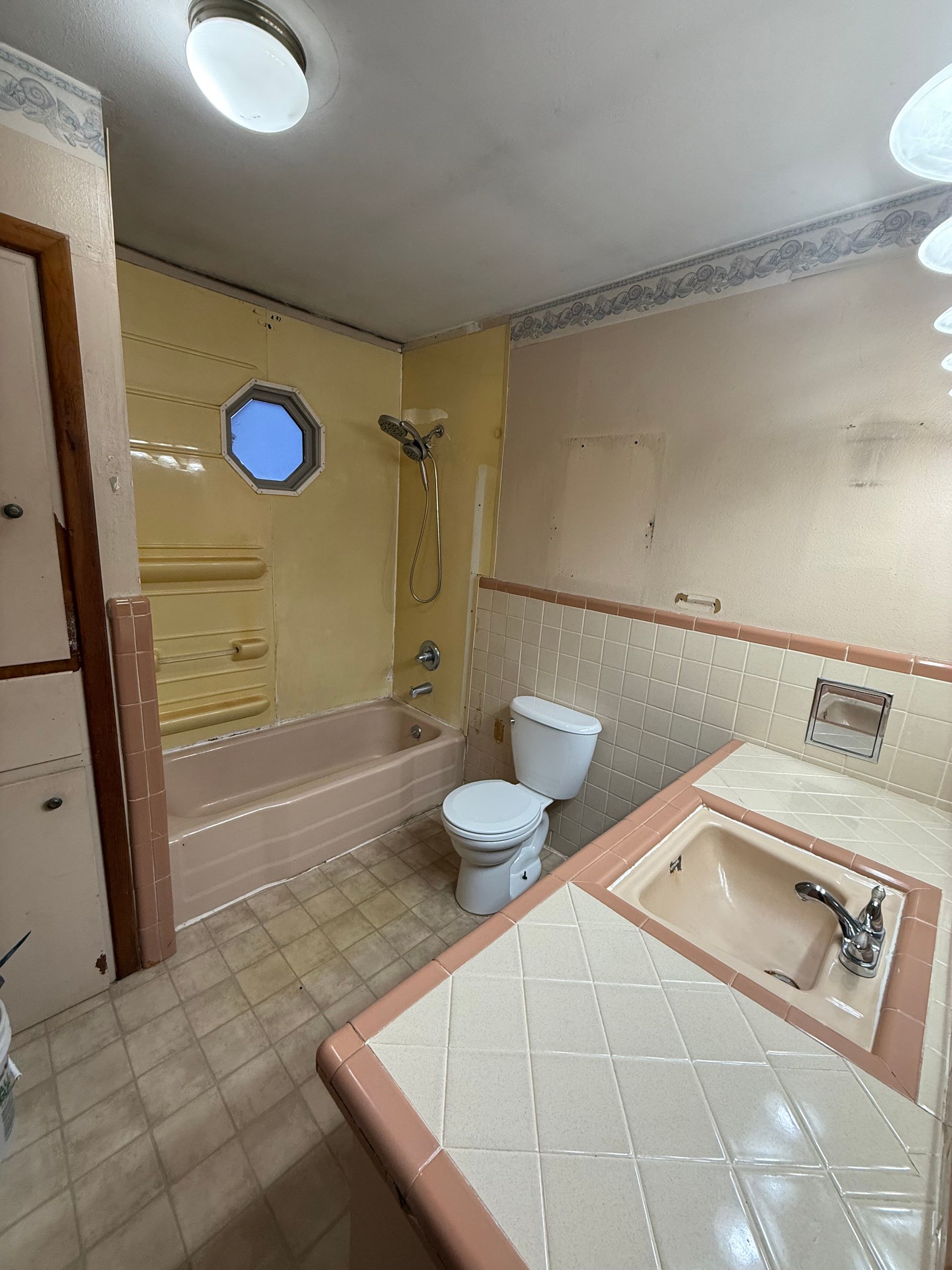 Small beige bathroom with pink tub, toilet, sink, round window, and tiled floor.
