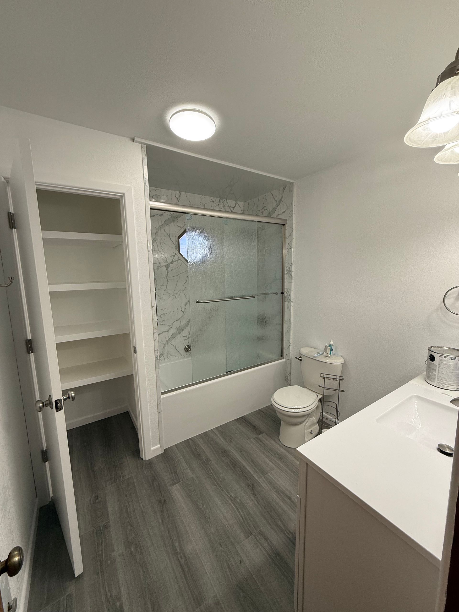 Small bathroom with white vanity, toilet, glass shower, open shelves, and gray wood-look floor.