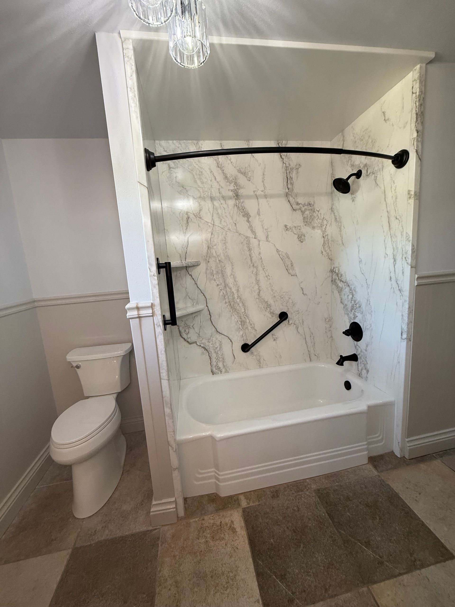 Accessible bathroom with white tub shower, marble wall surround, grab bars, toilet, and tiled floor