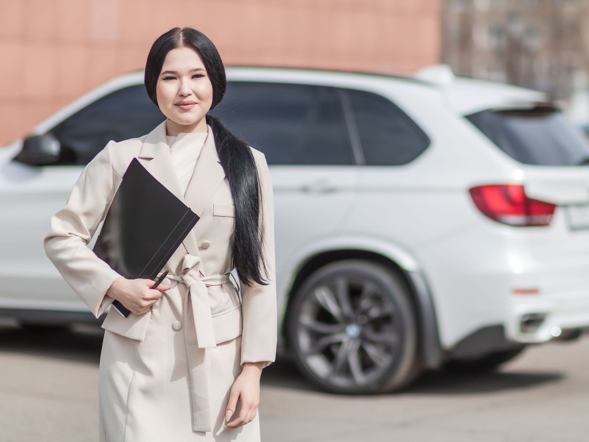 Woman in a beige suit holding a black folder, standing in front of a white SUV.