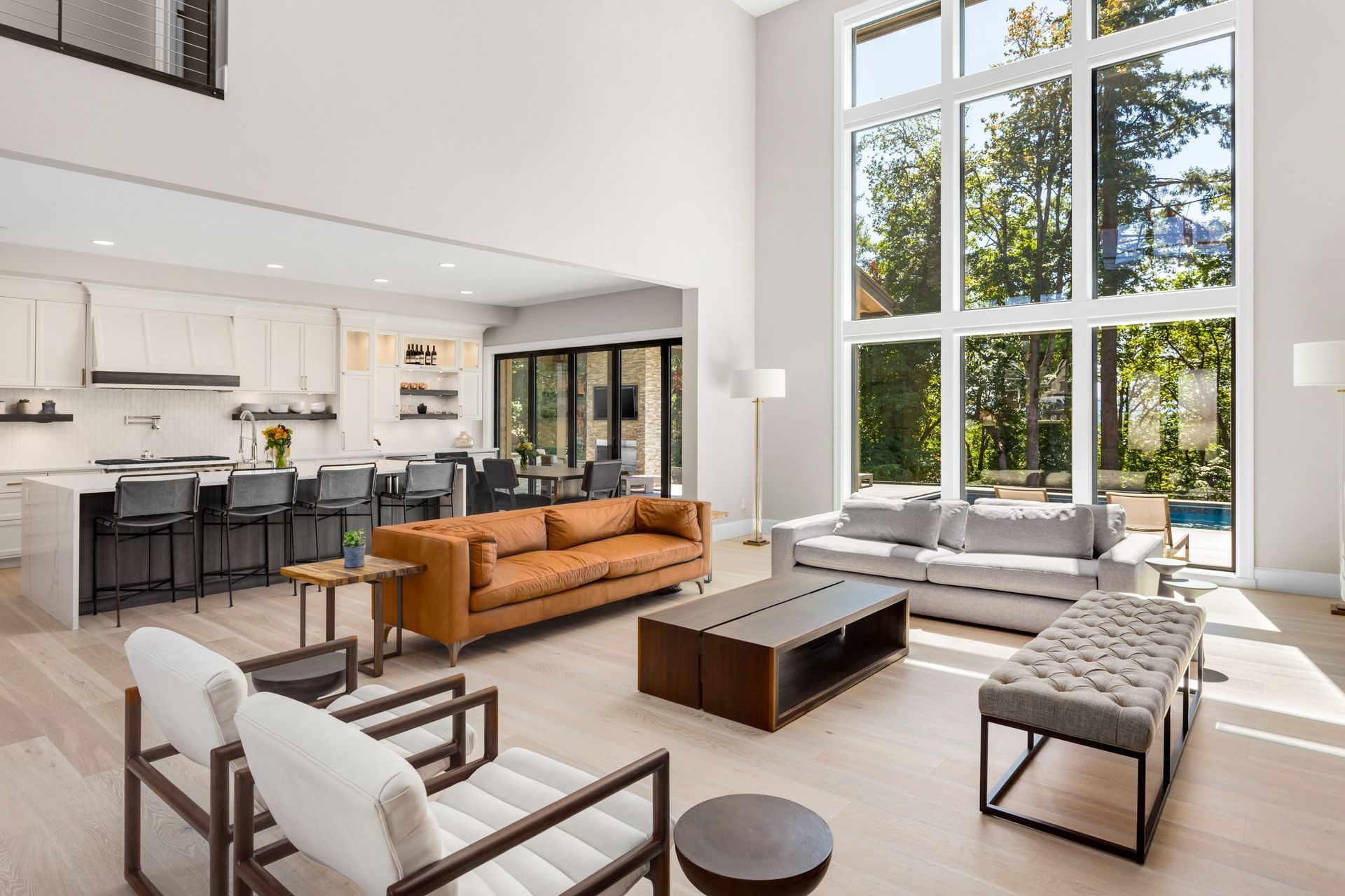 A sunlit, open-concept living room with a brown leather sofa, white chairs, a coffee table, and a modern kitchen.