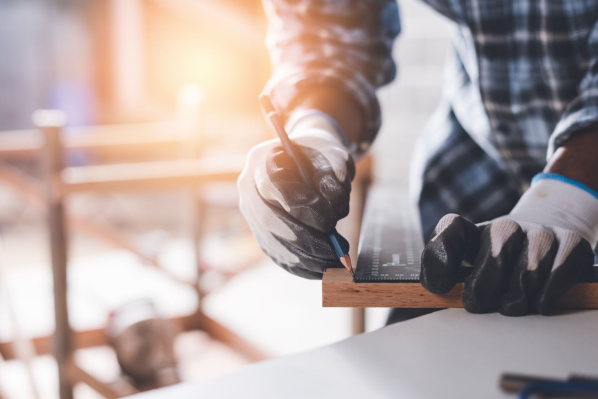 A person wearing work gloves marks a measurement on a wooden board with a pencil and a metal ruler. A person wearing work gloves marks a measurement on a wooden board with a pencil and a metal ruler.