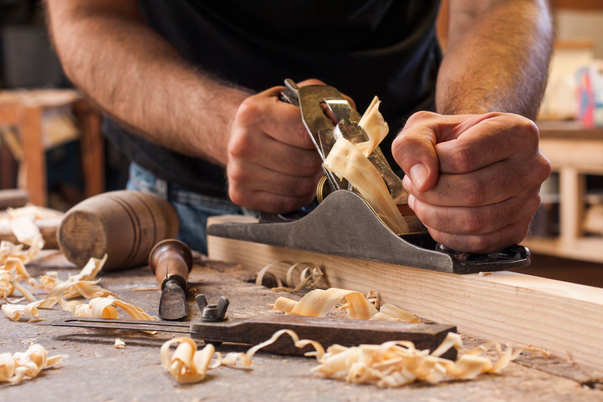 A person using a hand plane to smooth a wooden board on a workbench covered in wood shavings. A person using a hand plane to smooth a wooden board on a workbench covered in wood shavings.