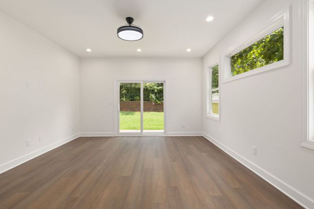 Empty modern room with white walls, wood floors, recessed lights, and large windows overlooking a green yard