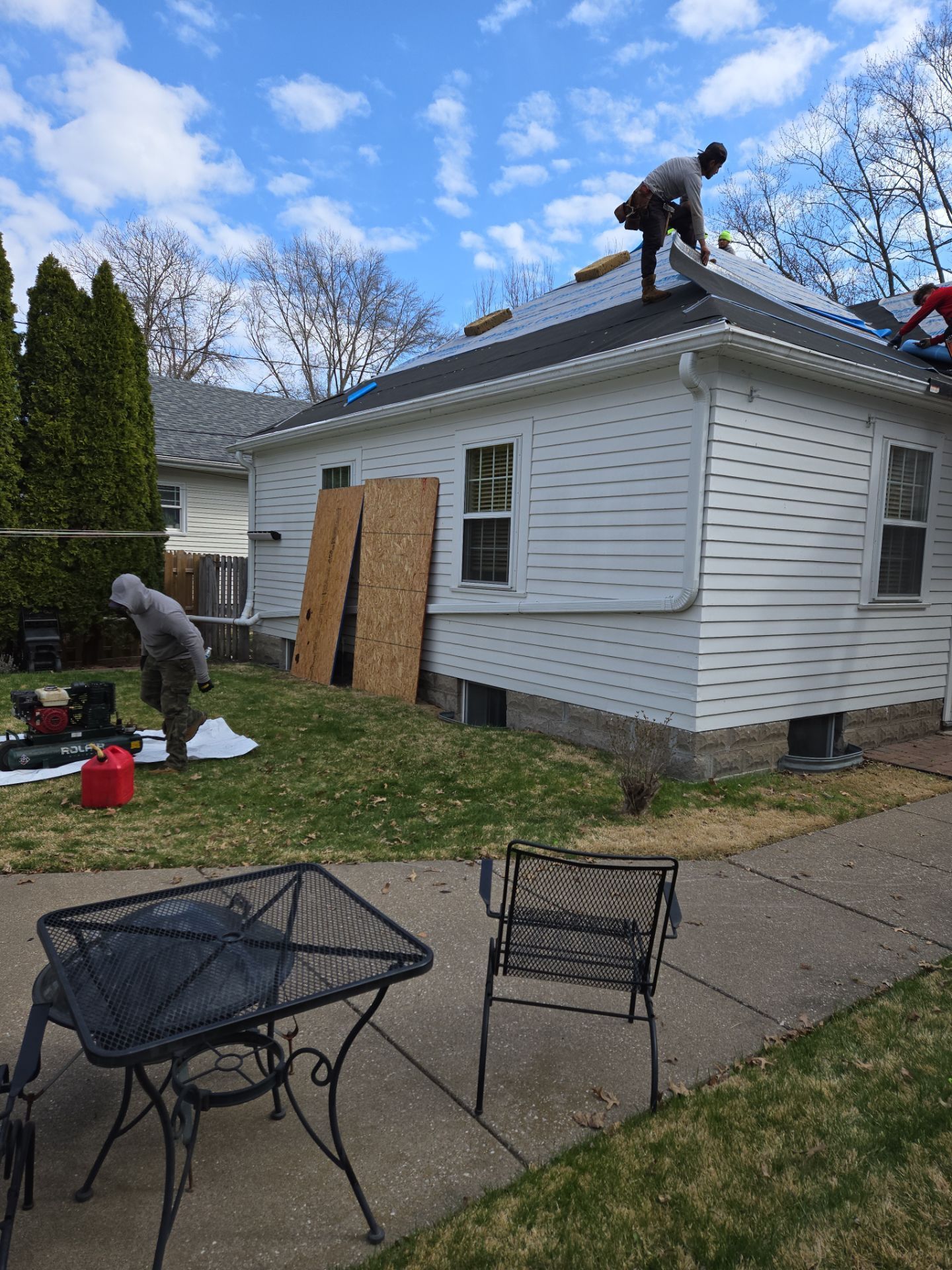 Workers repairing roof on a white house with plywood over a window and lawn tools in front