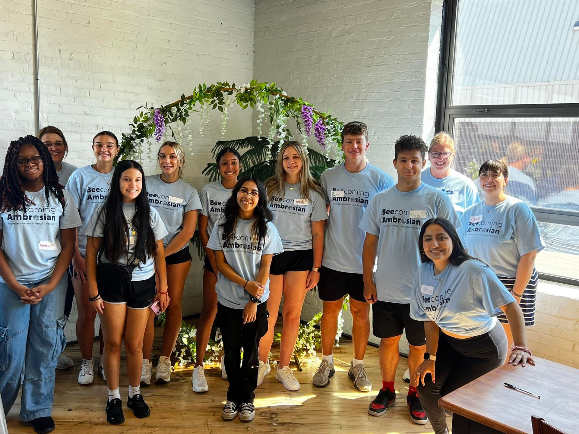 Group of students in light gray shirts posing indoors beneath a floral arch in a bright room.