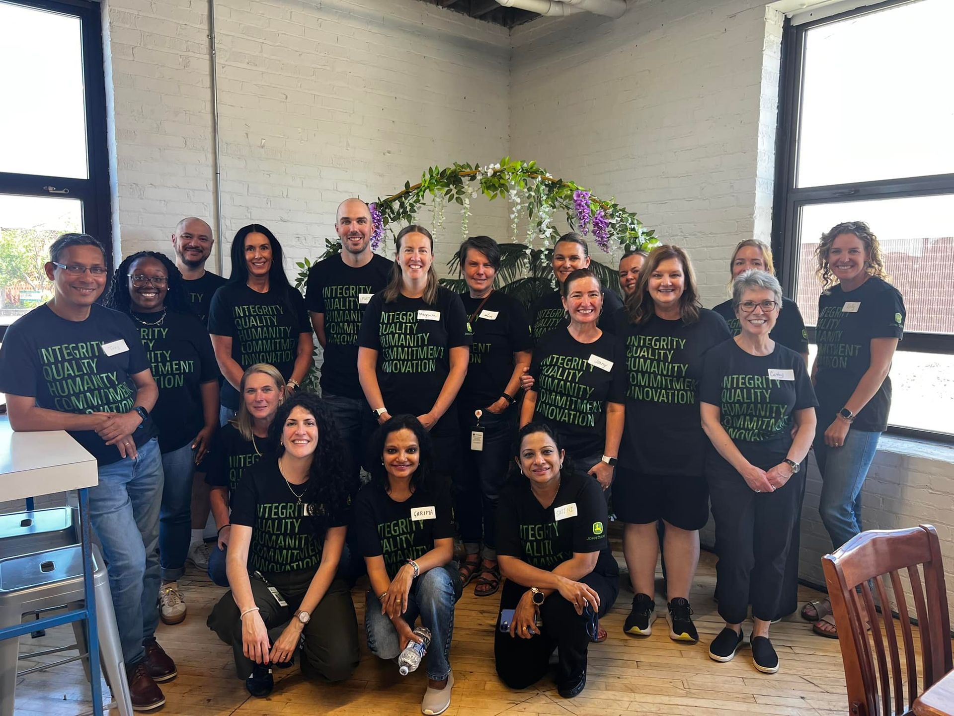 Group of people in matching black shirts posing indoors in front of a floral arch and large windows.