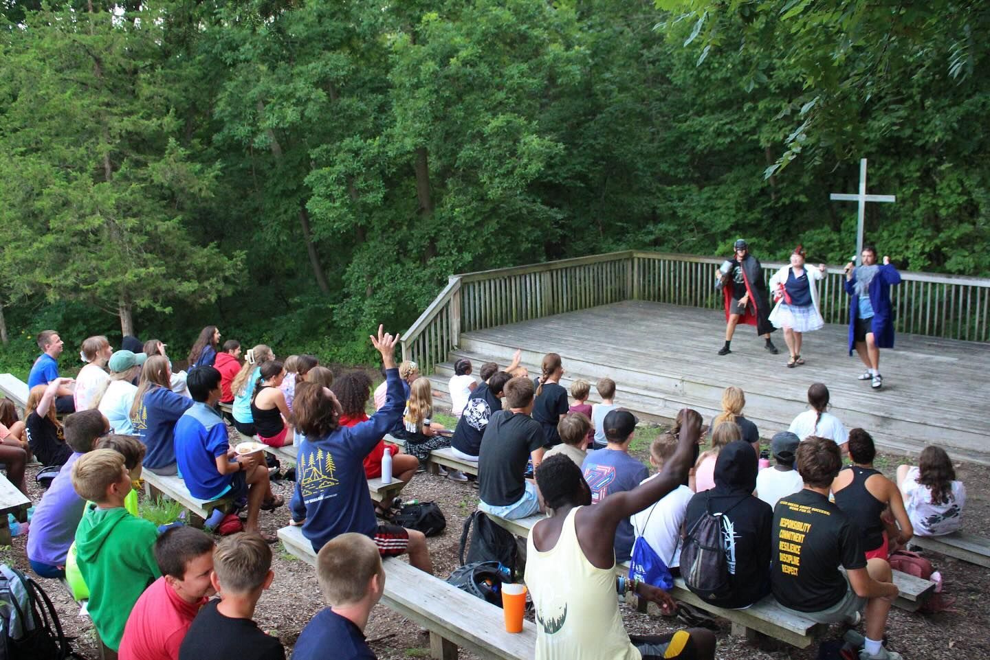 Outdoor worship service with people gathered on benches, facing a stage with a cross in a wooded setting