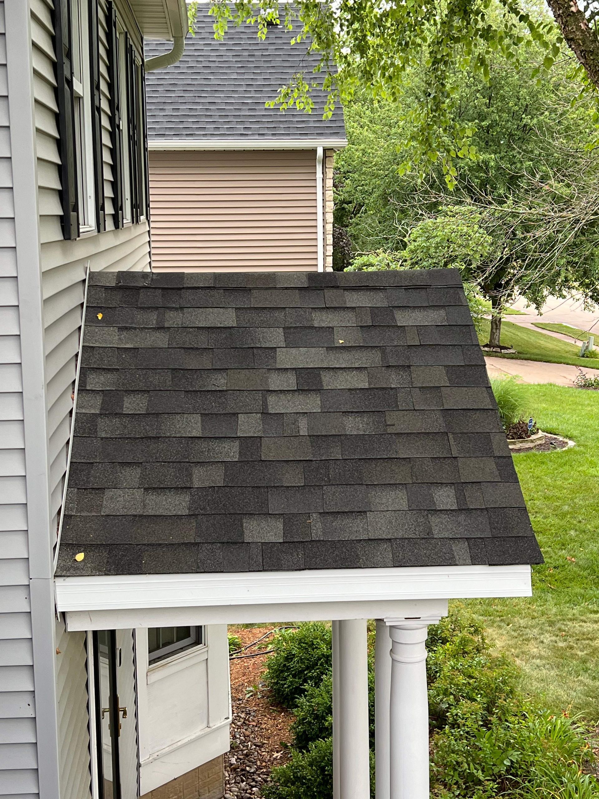 Small dark shingled porch roof attached to a house, with white trim and columns, beside a yard.