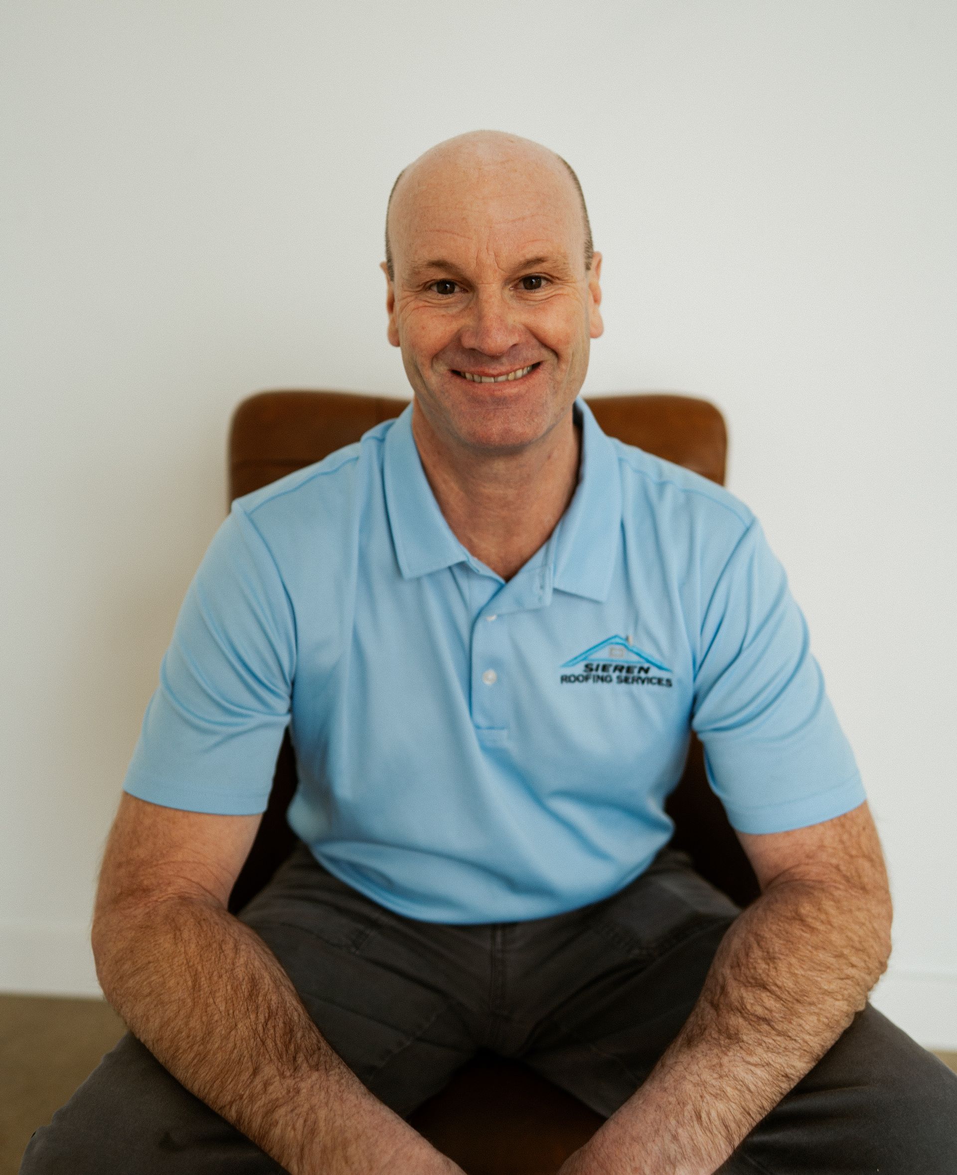 Smiling man in a light blue polo shirt seated on a chair against a plain white wall.