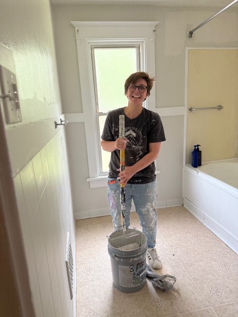 Person painting a bathroom wall white with a roller, standing beside a paint bucket and tray.