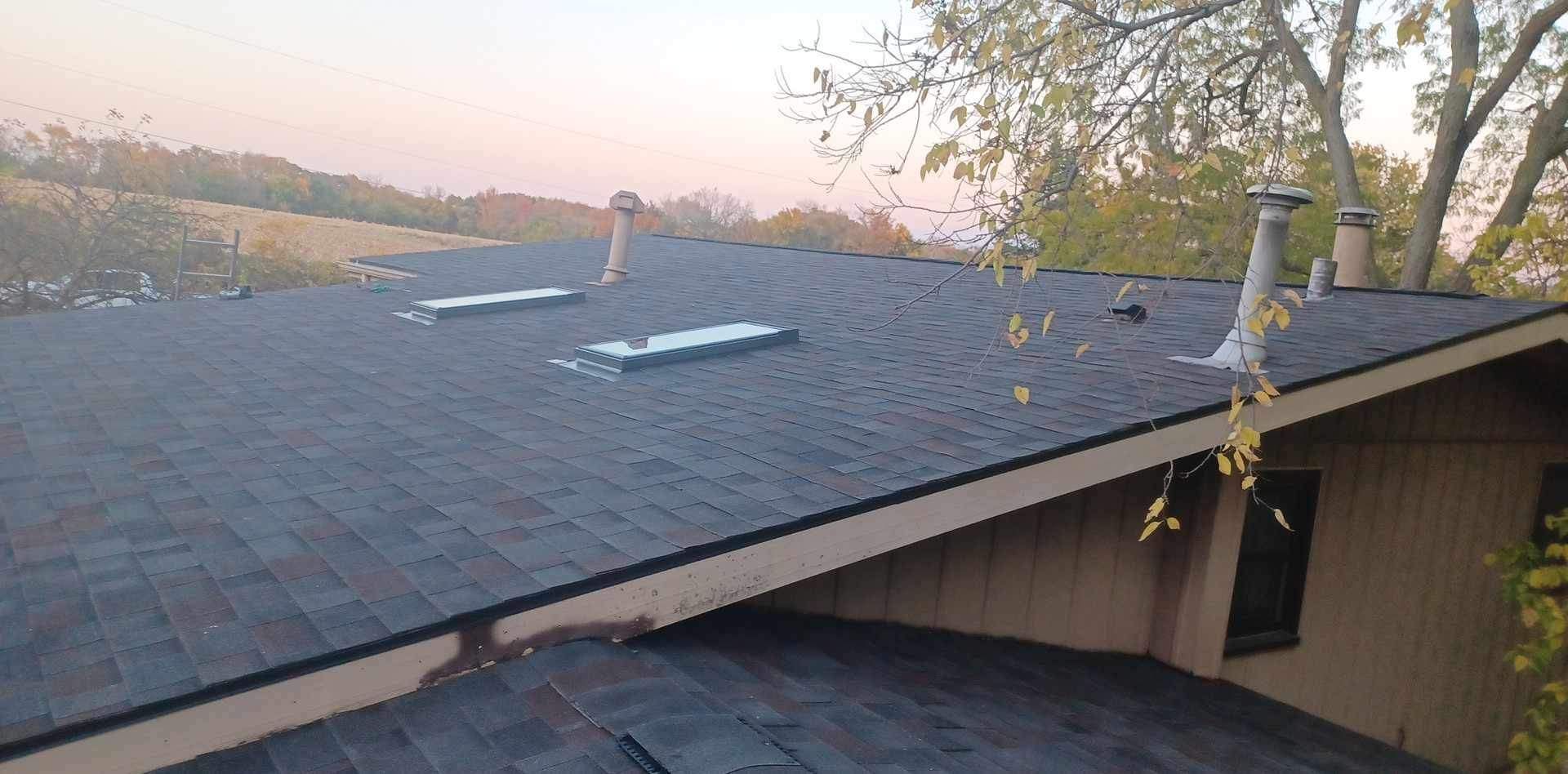 Residential roof with gray shingles and a skylight at sunset, viewed from above.