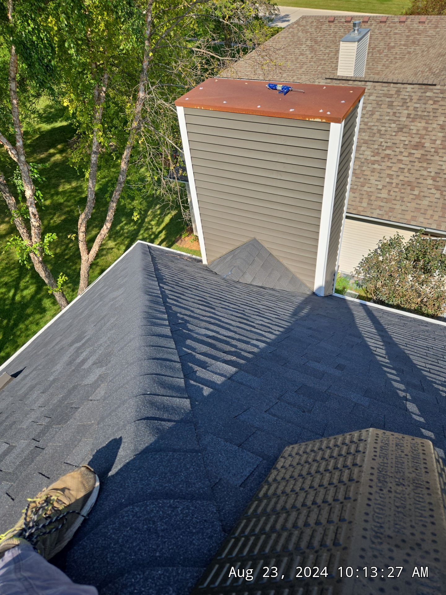 Rooftop view of a house chimney beside trees, with dark gray shingles and a small brown cap on top.