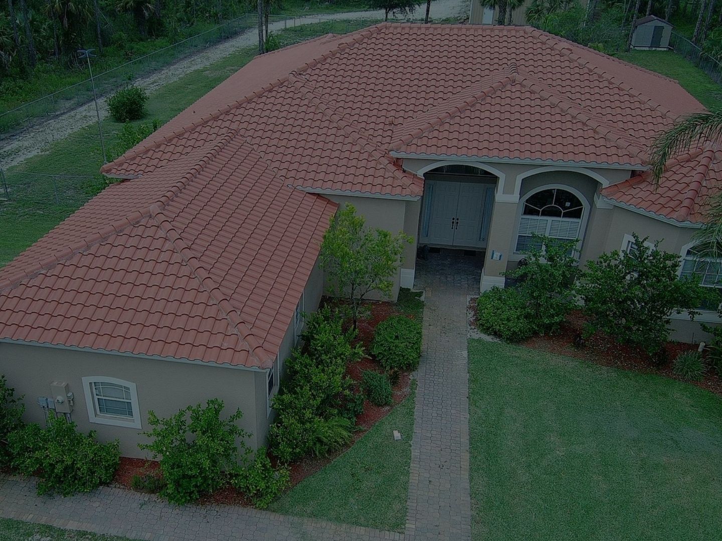 Aerial view of a tan house with a red tile roof and front walkway, surrounded by green lawn and shrubs.