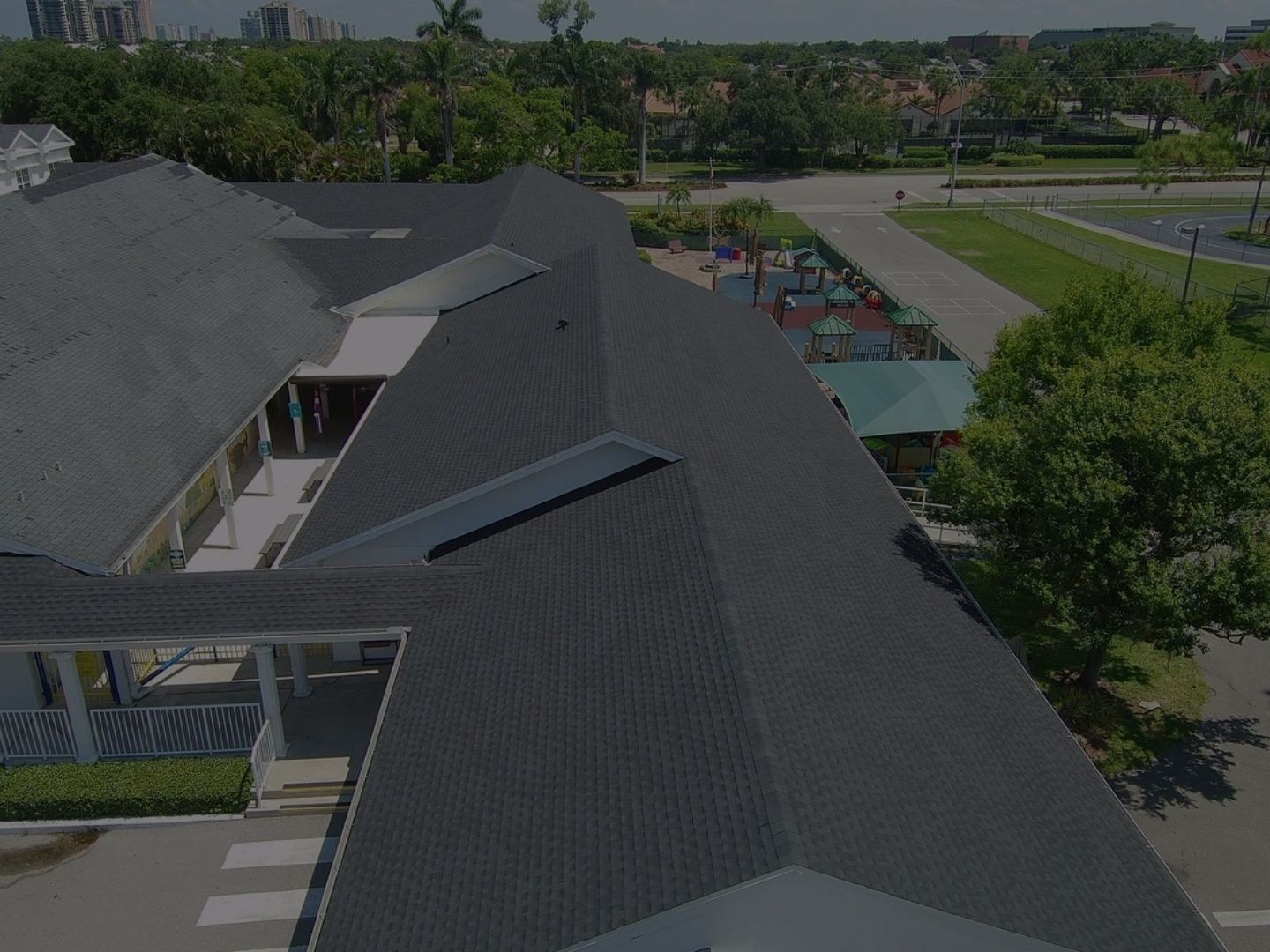Aerial view of a dark-roofed building beside a roadway and trees, with a small playground in the distance