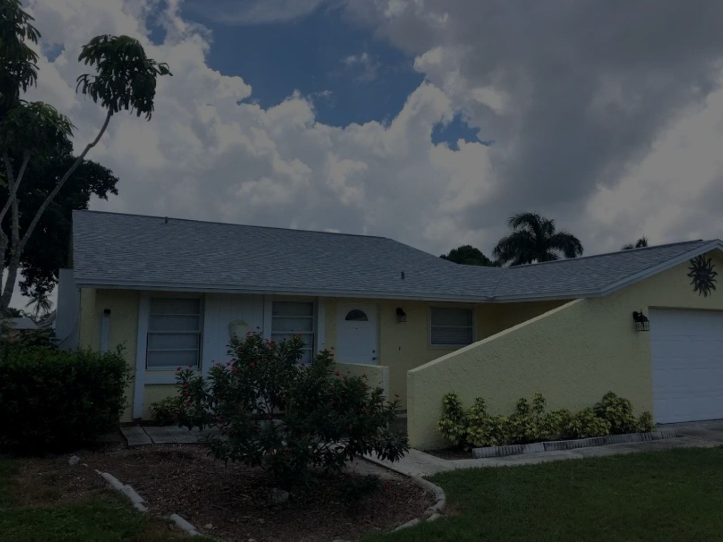 Single-story house with gray roof, pale yellow walls, and a white garage under a cloudy sky