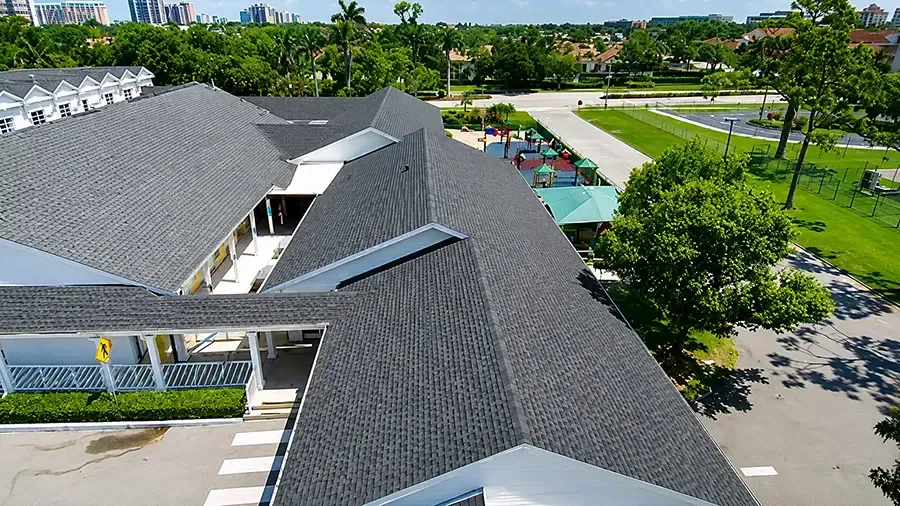 Aerial view of a gray-roofed building beside a tree-lined parking lot and road