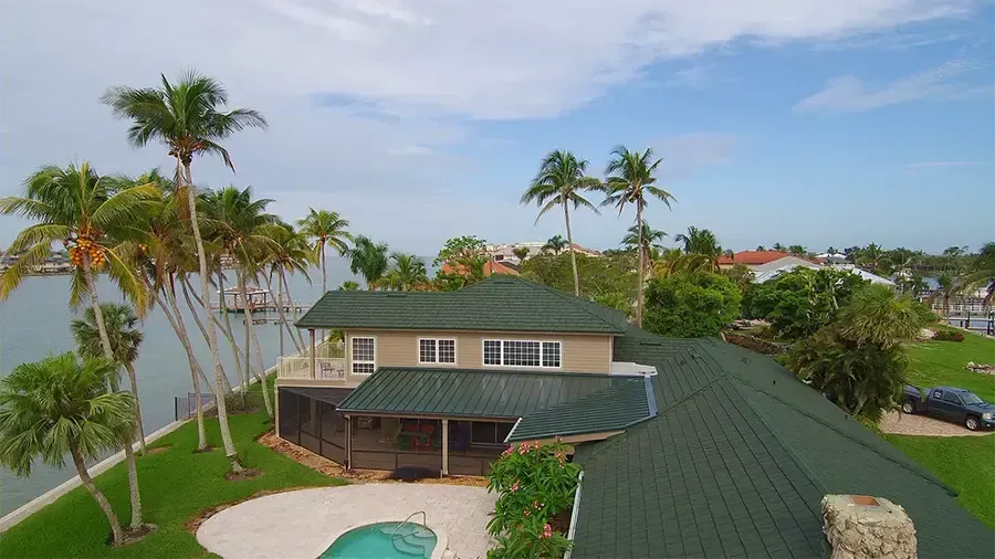 Aerial view of a tropical house with green roof, palm trees, and a small backyard pool near the coast