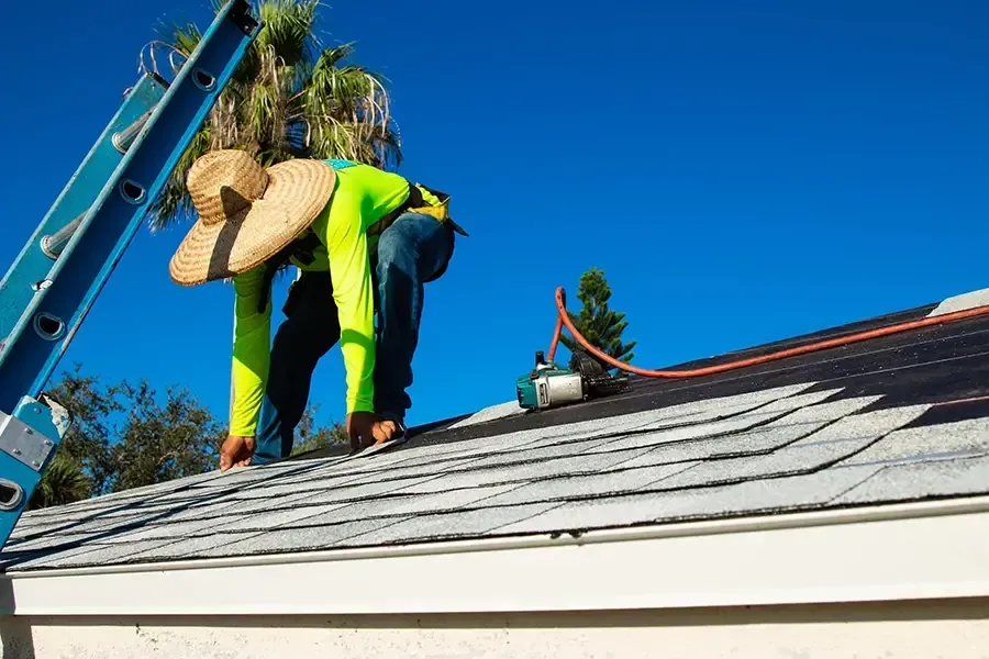 Workers on a roof near a blue ladder, inspecting shingles under a clear blue sky