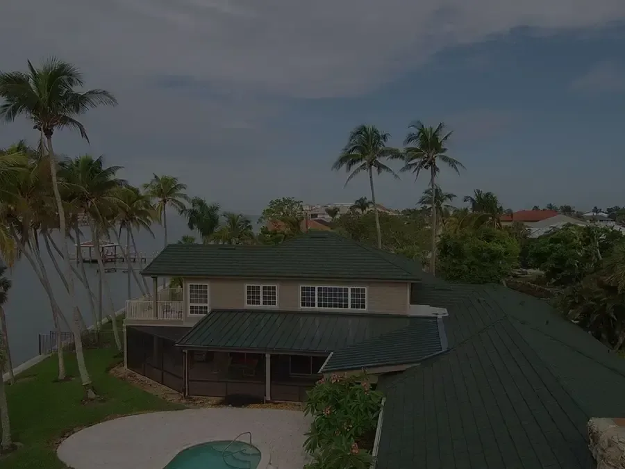 Aerial view of a dark green-roofed house beside palm trees and water at dusk.