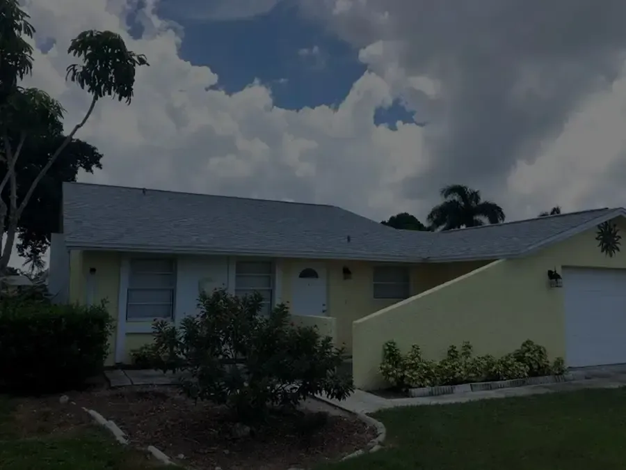 Single-story suburban house with gray roof, pale yellow walls, and cloudy sky overhead
