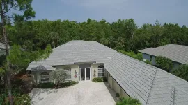 Stone house with a gray shingled roof, white driveway, and trees surrounding it under a blue sky