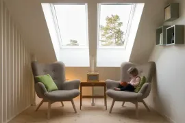 Bright attic room with two armchairs, a child reading on one chair, and skylights above the desk.