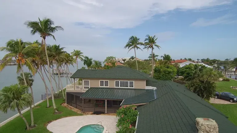 Aerial view of a tropical house with palm trees, a green roof, and a small backyard pool near the coast.