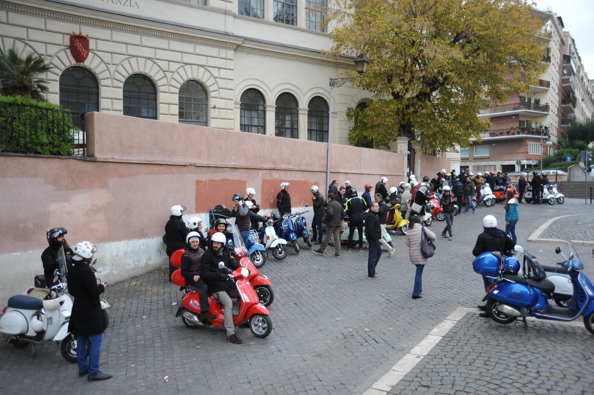 Motorcyclists in helmets line up on a city street beside a stone building and pink wall