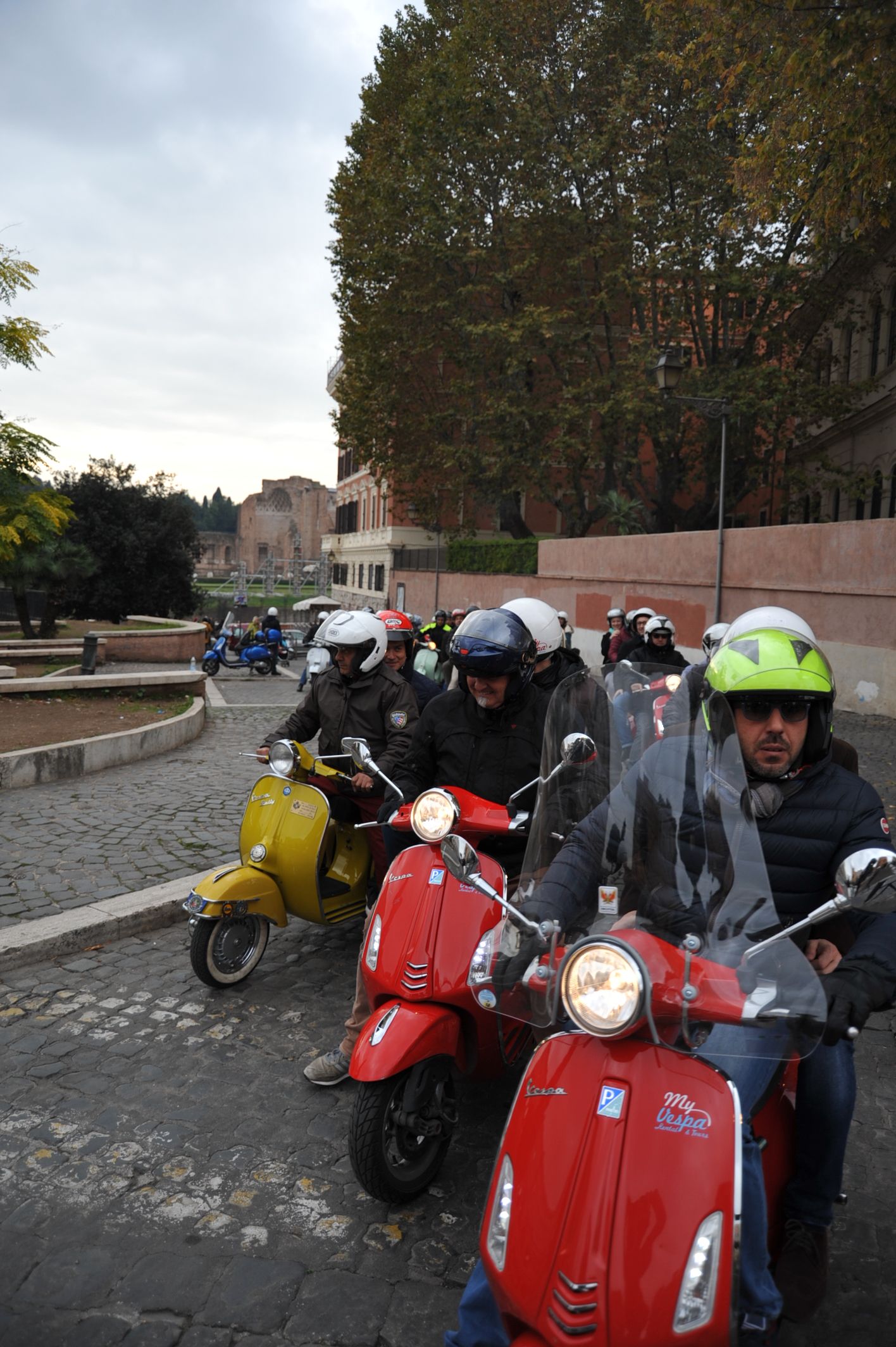 Scooters lined up on a wet street, riders wearing helmets in the foreground.