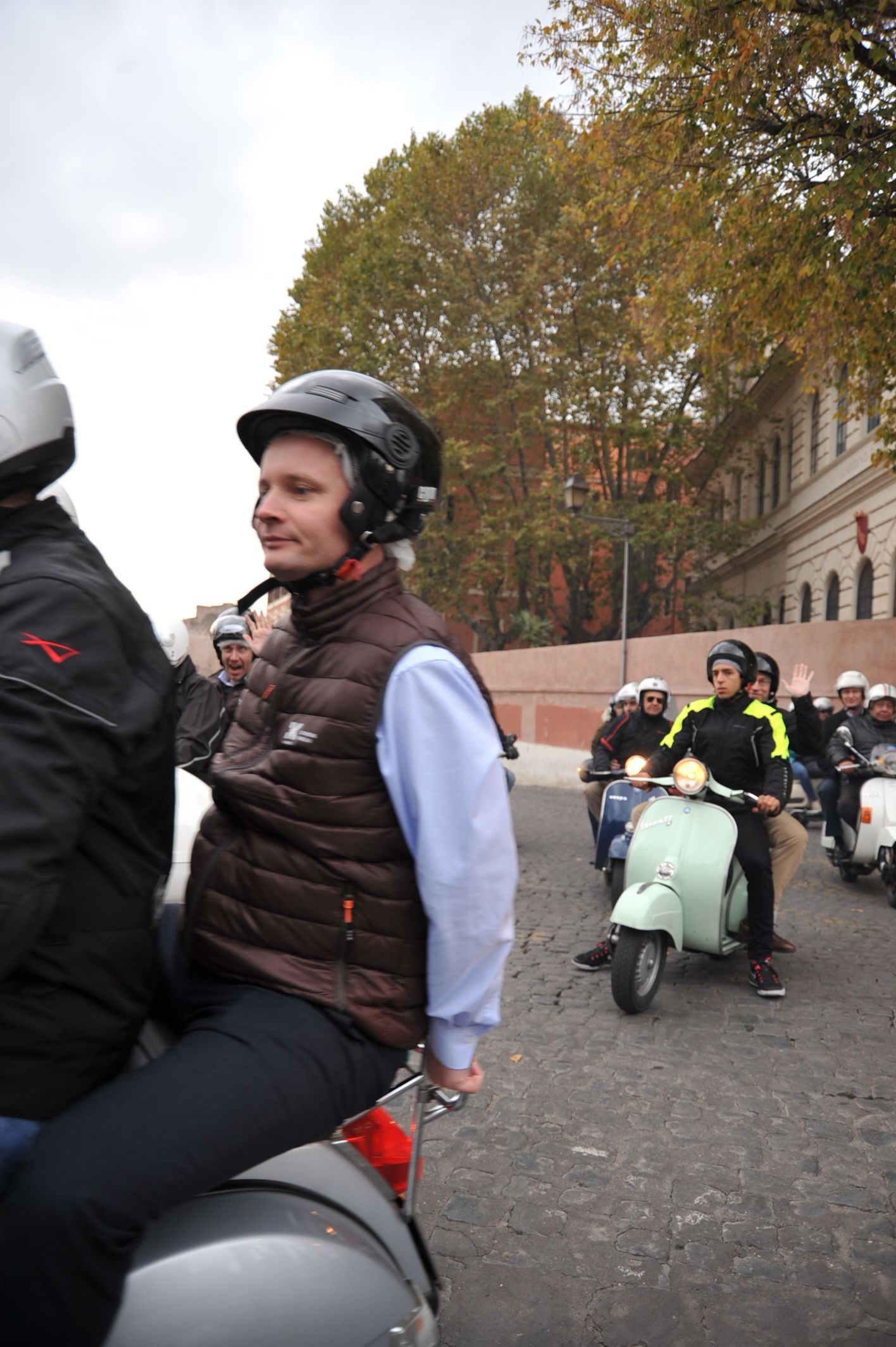 People riding scooters on a city street, one in a helmet and black jacket in the foreground