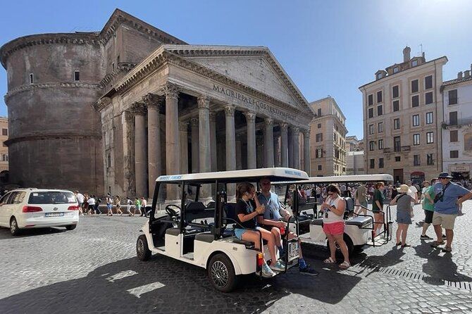 Tourists ride a white open shuttle past a historic stone temple and cobblestone square on a sunny day