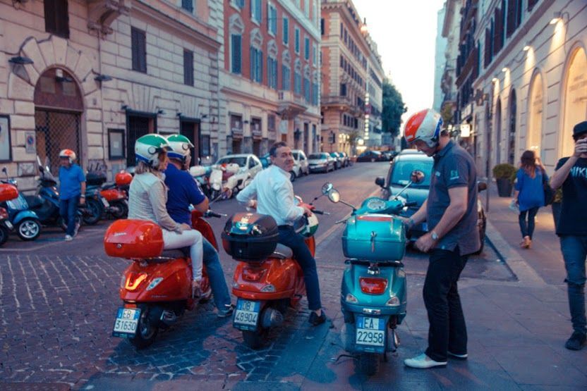 People on colorful scooters gathered on a cobblestone city street between tall buildings.