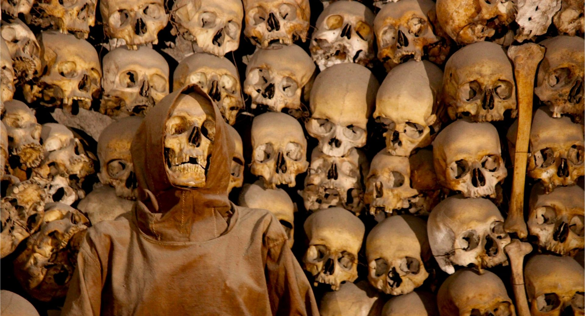 Rows of human skulls and bones stacked behind a mummified body in a dark ossuary display