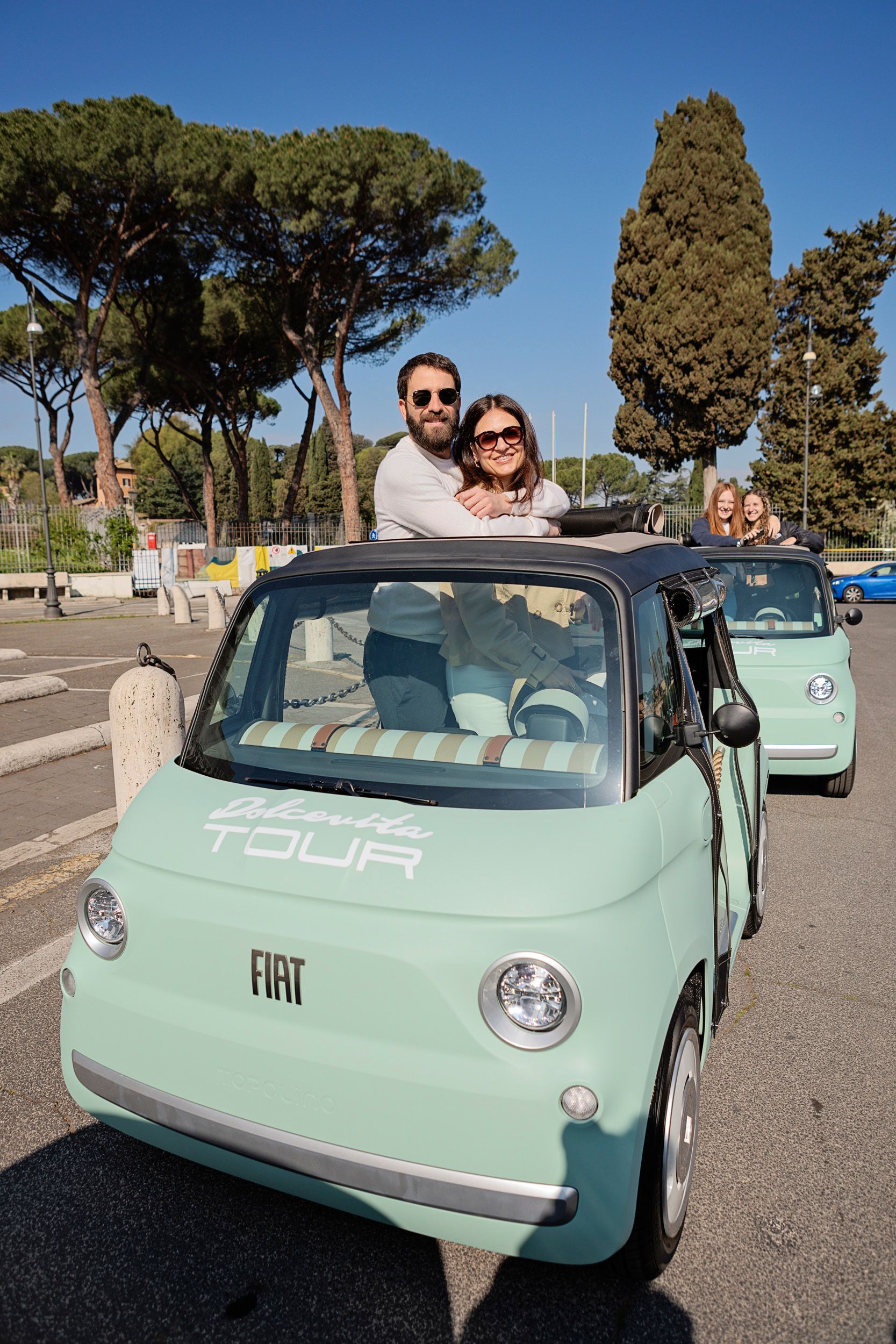 Two people posing in a mint-green vintage car on a sunny street with trees and parked cars.
